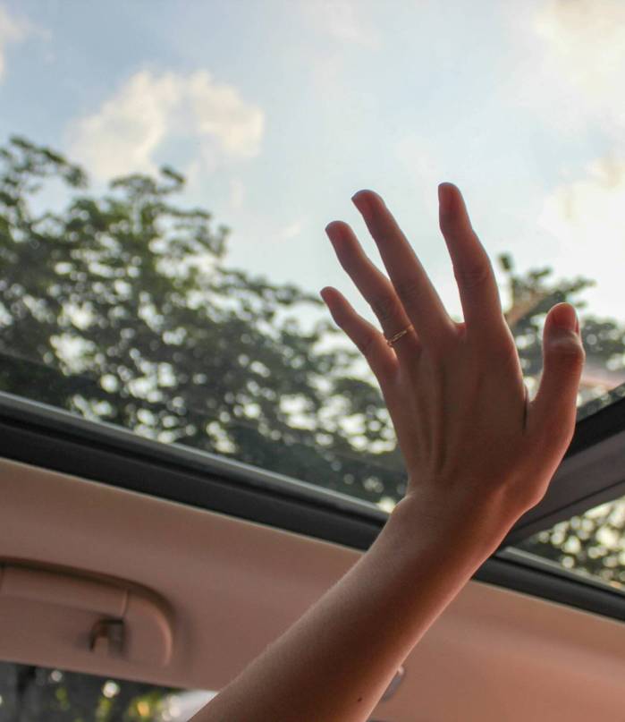 A hand reaching out through a car sunroof, touching the sky on a sunny day.
